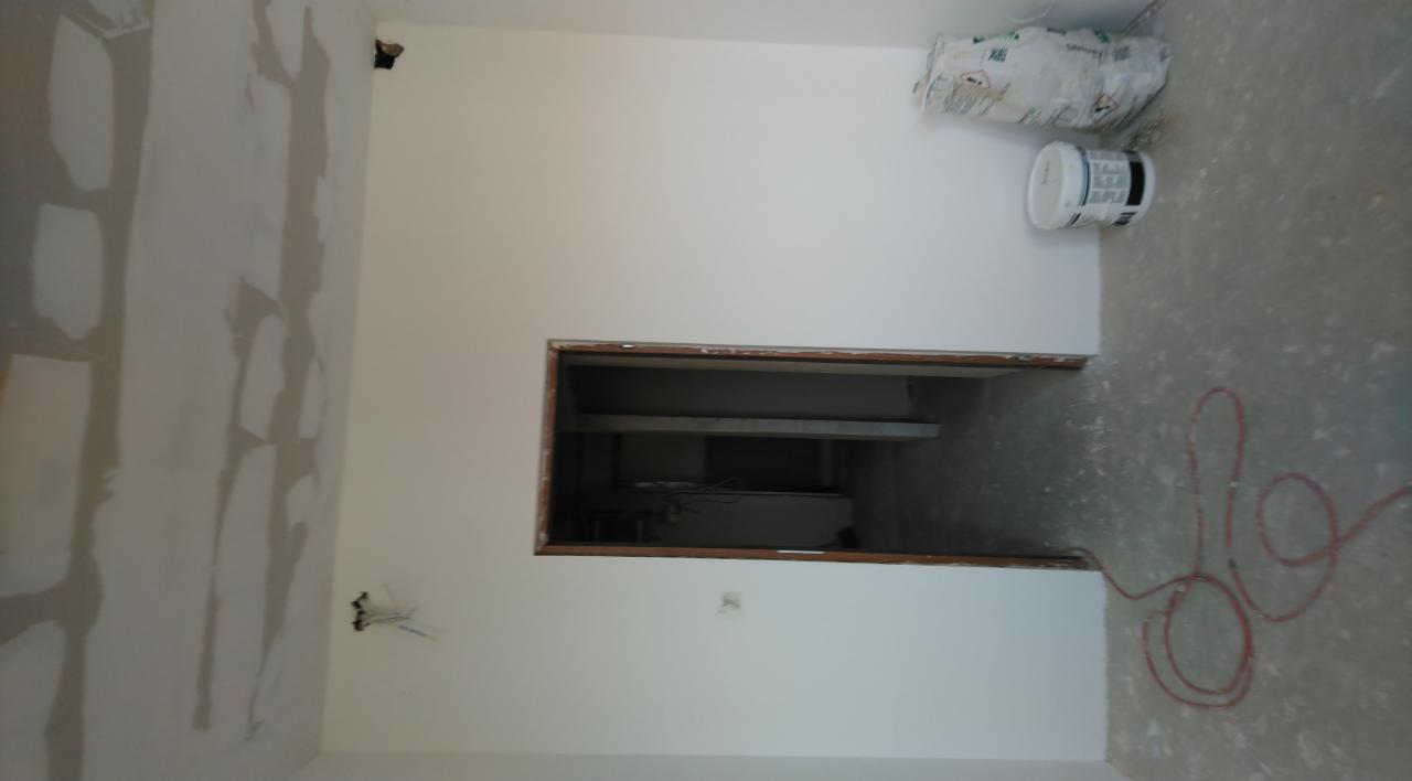 Wnętrze w trakcie remontu: ściana z częściowo nałożoną warstwą szpachli, bęben z farbą, worek z materiałem budowlanym i wystające przewody elektryczne.