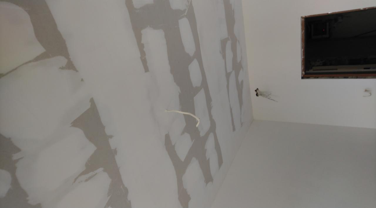 Ściana połatana gładzią szpachlową, widoczne ślady łączeń płyt gipsowo-kartonowych przed szlifowaniem, wystające przewody elektryczne i otwór okienny w górnym rogu.