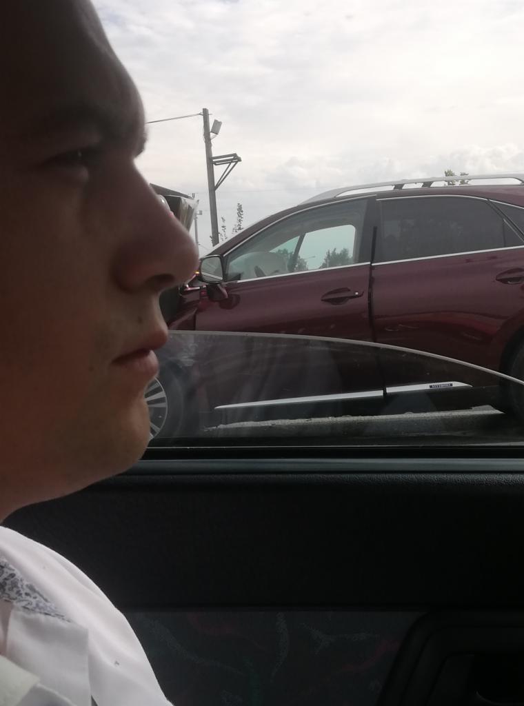 Profil mężczyzny w samochodzie, z widokiem na zaparkowany, bordowy SUV i słup energetyczny na tle pochmurnego nieba.