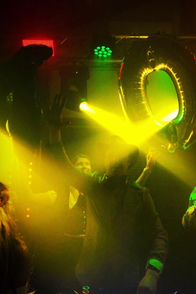 Osoby na parkiecie w stroboskopowym oświetleniu, dekoracja w postaci balonu w kształcie cyfry zero, widoczne elementy nagłośnienia i oświetlenia scenicznego.