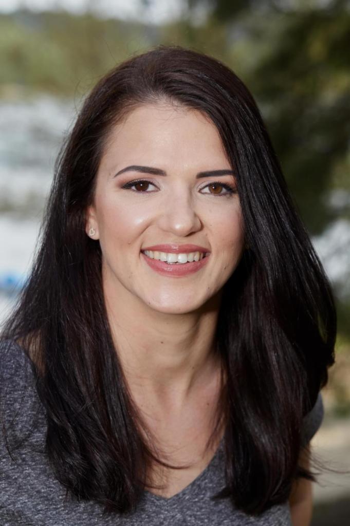 Portret uśmiechniętej brunetki z ciemnymi włosami i subtelnym makijażem, na rozmytym, zielonym tle. Widoczne delikatne kolczyki.