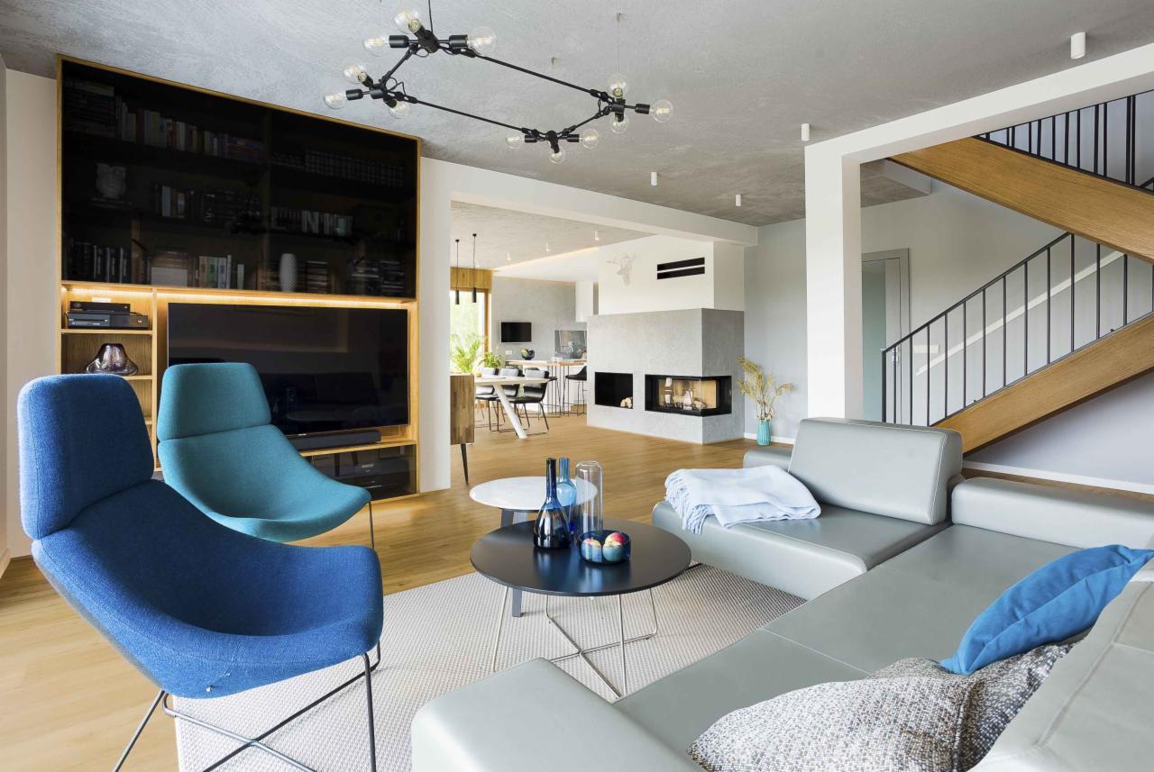Nowoczesny salon z szarym narożnikiem, dwoma fotelami w odcieniach niebieskiego, kominkiem i schodami z drewnianymi stopniami, oświetlony designerską lampą sufitową.