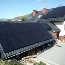 Czarny panel słoneczny na ciemnym dachu dwuspadowym domu jednorodzinnego z kominem z czerwonej cegły, widok z góry na podwórze z kostki brukowej i trampoliną.