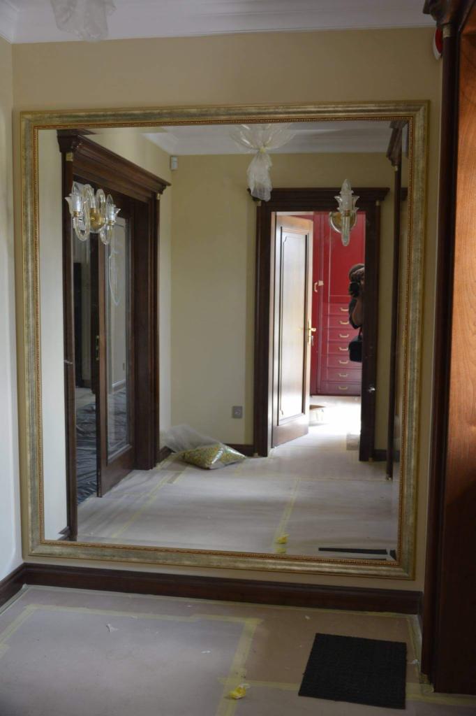 Duże lustro w złotej ramie zawieszone na ścianie w korytarzu, widoczne zabezpieczenia podłogi podczas montażu.