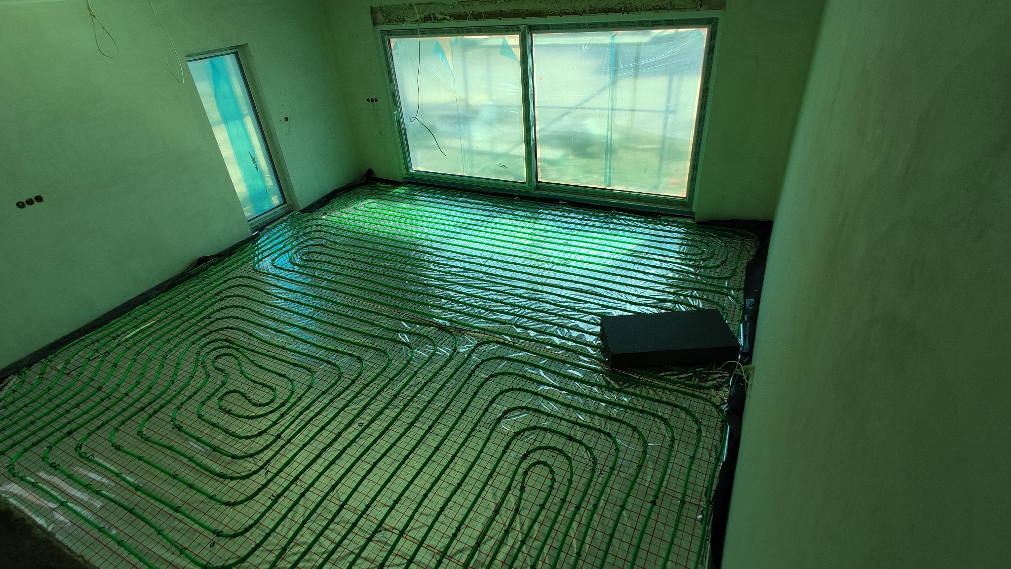 Zielone rury ogrzewania podłogowego ułożone na folii w surowym wnętrzu z oknami zabezpieczonymi folią malarską.