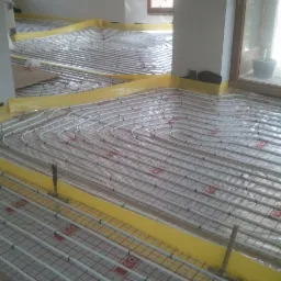 Białe rury ogrzewania podłogowego ułożone na folii z czerwoną kratką, zabezpieczone żółtą taśmą przy ścianach w trakcie montażu w jasnym pomieszczeniu.