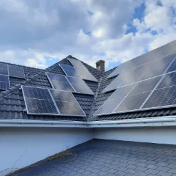 Panele słoneczne zamontowane na dachu domu jednorodzinnego z ciemną dachówką i ceglanym kominem, widok z dołu na tle pochmurnego nieba.