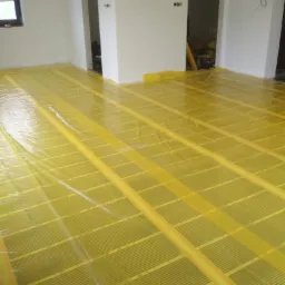 Żółta folia grzewcza rozłożona na podłodze w surowym stanie, przygotowana do montażu, widoczne puste otwory drzwiowe i fragment okna.