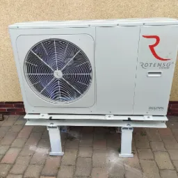 Biała zewnętrzna jednostka pompy ciepła marki Rotenso Aquami, zamontowana na metalowej konstrukcji wsporczej, na tle beżowej ściany i kostki brukowej. Widoczne logo producenta.