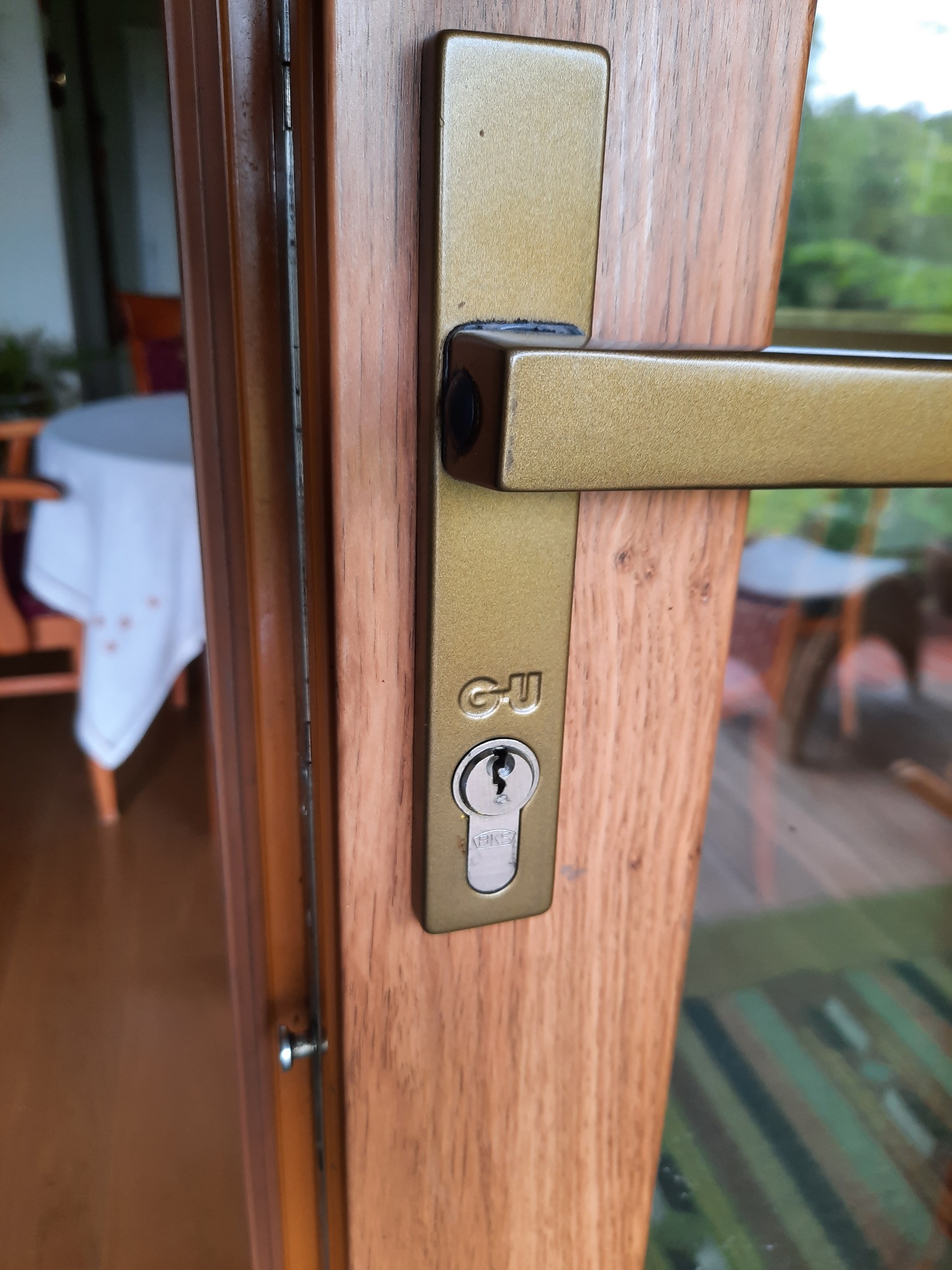 Złota klamka okienna z zamkiem na drewnianej ramie, widoczne logo 'GU', w tle rozmyty widok wnętrza z białym obrusem na stole.