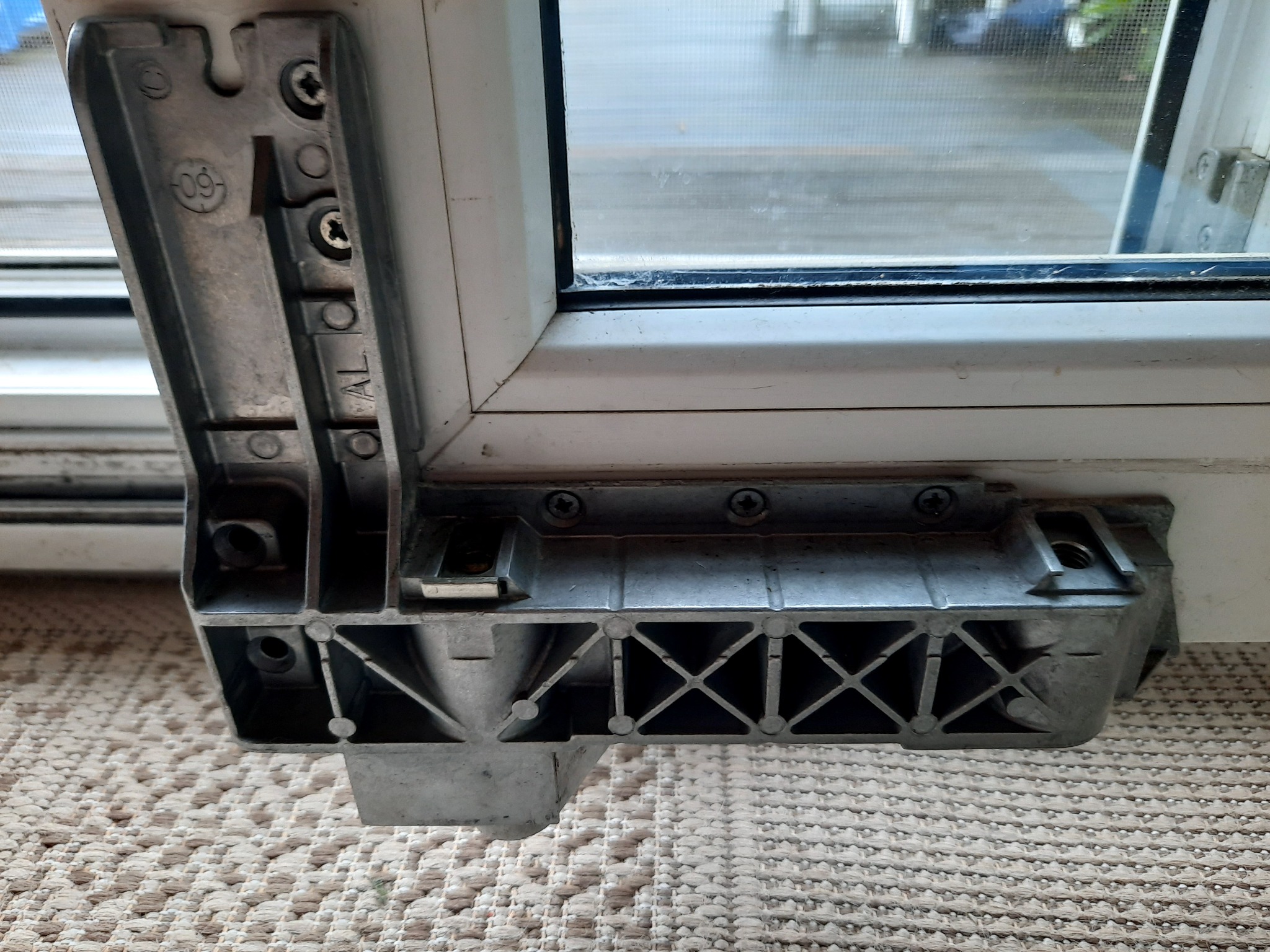 Szczegół mechanizmu okiennego, widoczne śruby mocujące i aluminiowy element z oznaczeniem '09 AL', biała rama okna w tle.