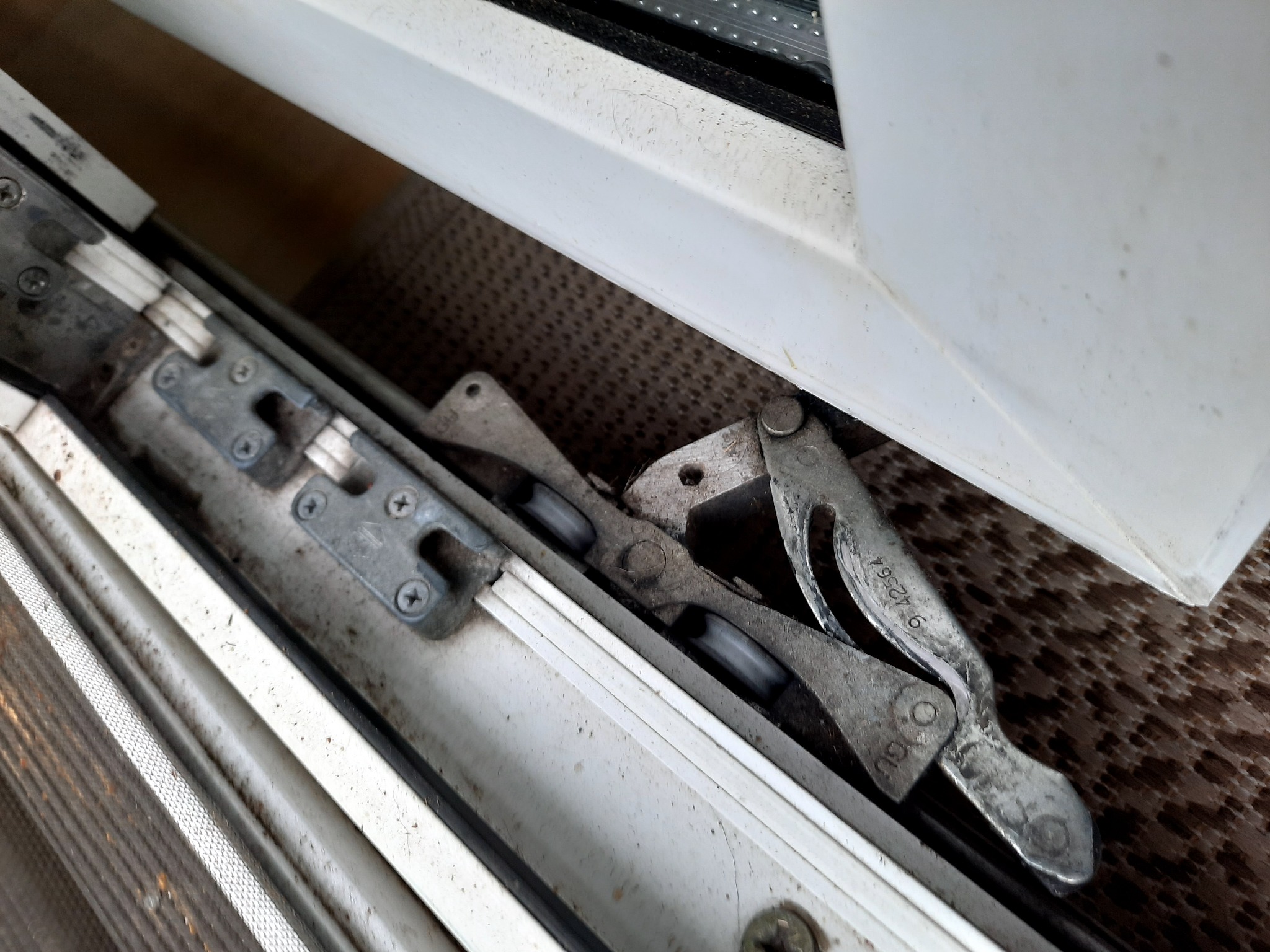 Detail metalowego mechanizmu okna z widocznymi śrubami i numerem seryjnym na ramieniu, ujęcie z bliska ukazuje zużycie i zabrudzenia.