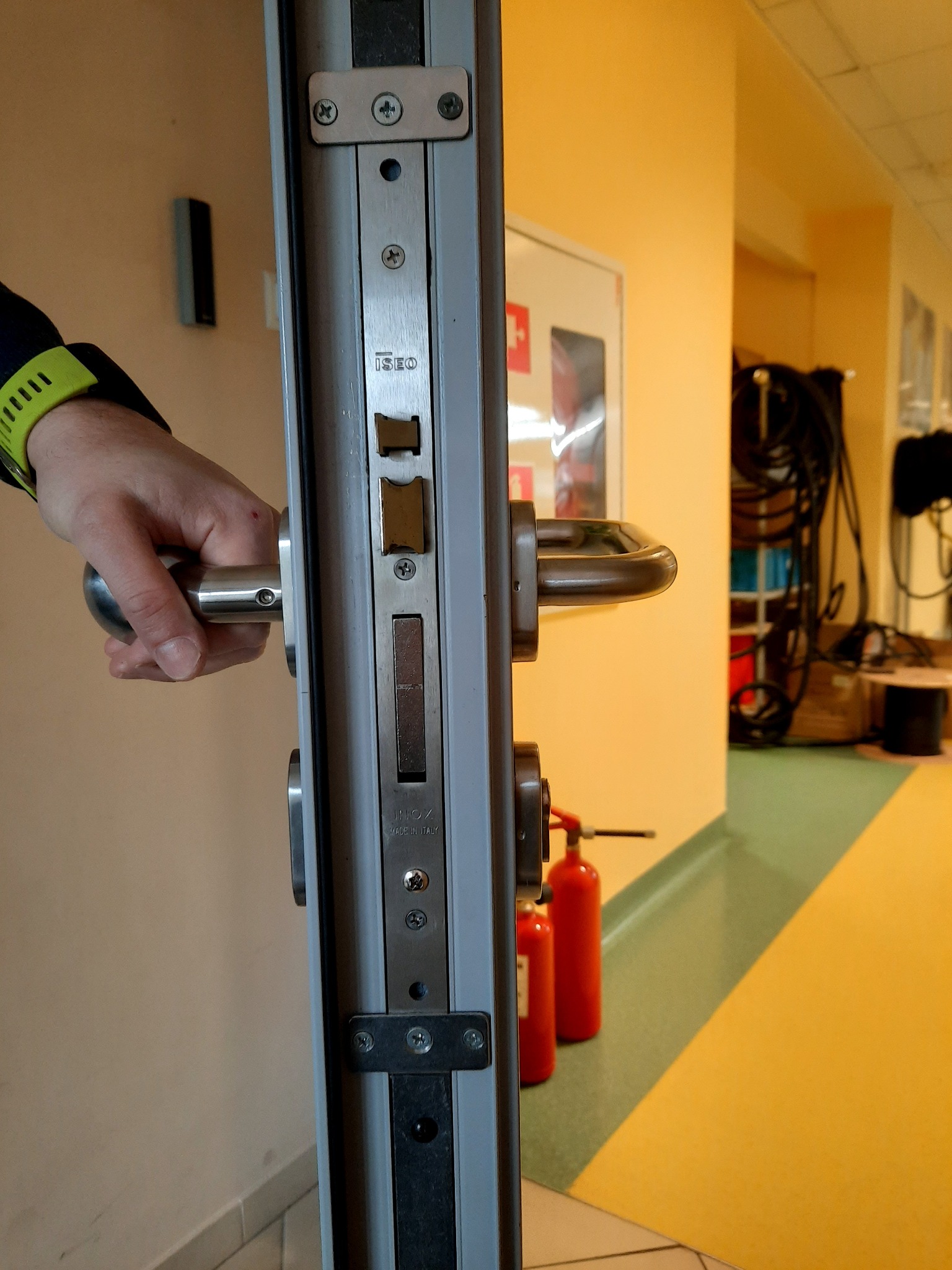 Otwieranie metalowych drzwi z zamkiem ISEO, widoczne dwa czerwone gaśnice w tle, wnętrze budynku.
