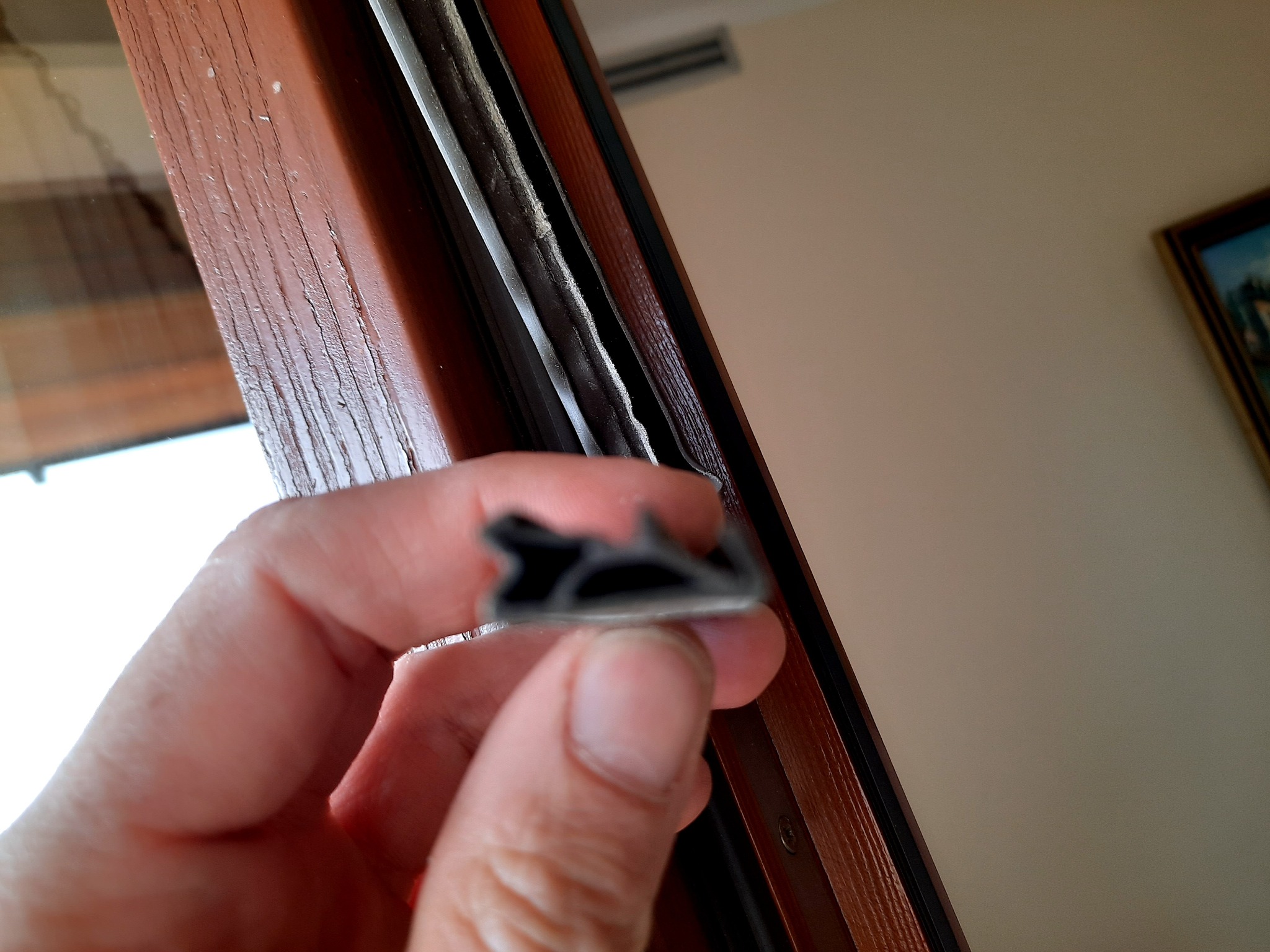 Dłoń trzyma profil uszczelniający okna na tle drewnianej ramy okiennej z widoczną szczeliną.
