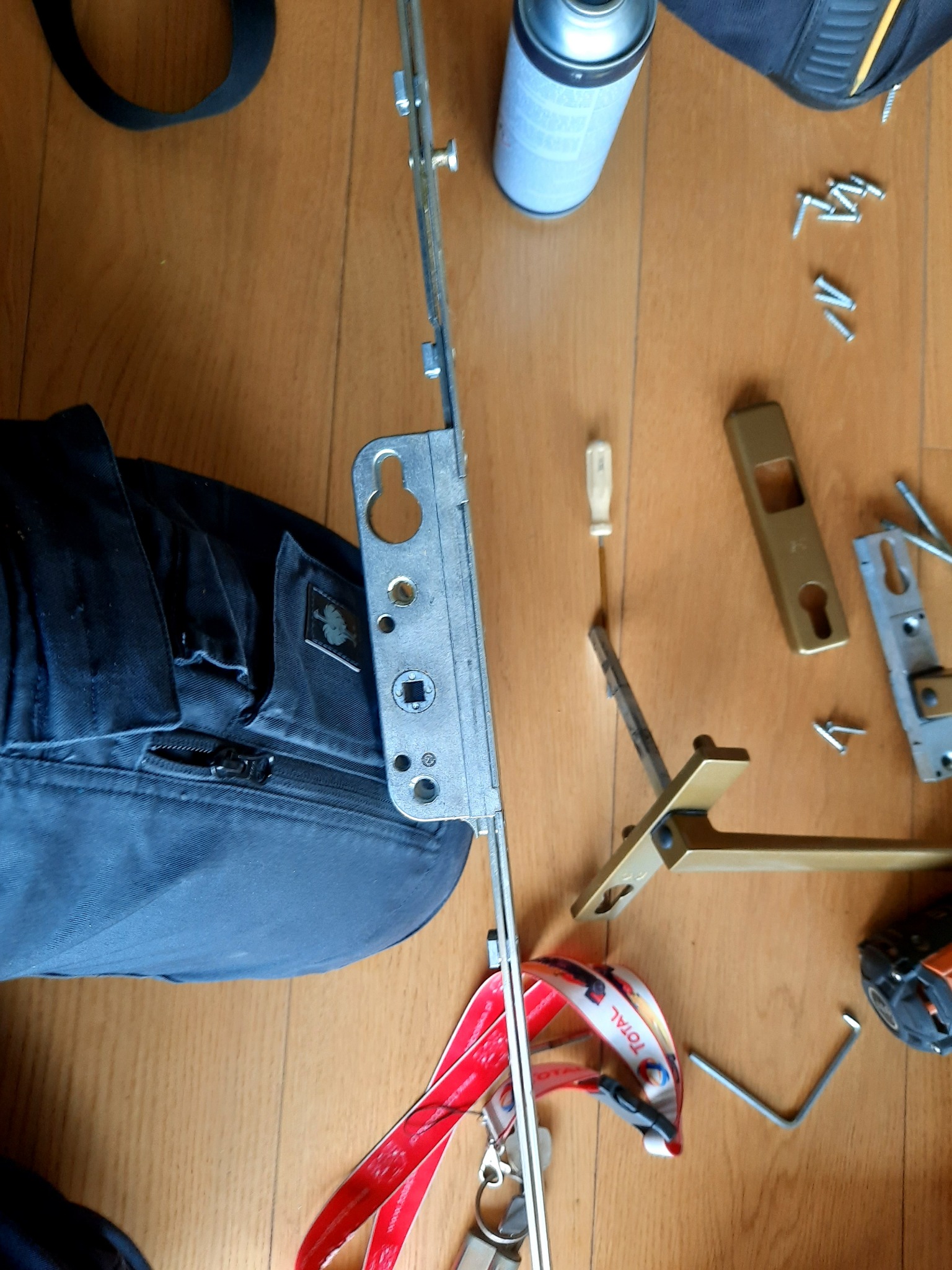 Rozłożony mechanizm zamka okiennego, wkrętak, puszka smaru, śruby i narzędzia na drewnianej podłodze obok torby narzędziowej z logo Engelbert Strauss.