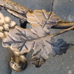 Detal metaloplastyki: dekoracyjny element w kształcie liścia i kiści winogron, w kolorach starego złota i czerni, na tle szarej kostki brukowej.