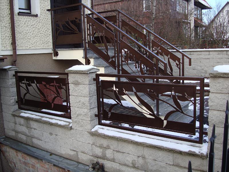 Metalowe ogrodzenie z motywem roślinnym i balustrada schodowa, wykonane w Wąbrzeźnie, z lekkim śniegiem na elementach kamiennych.