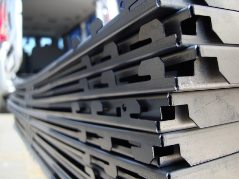 Stos wygiętych, metalowych profili z charakterystycznymi wycięciami, prawdopodobnie elementów konstrukcyjnych ogrodzenia panelowego, ułożonych wewnątrz pojazdu.