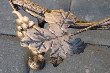 Detal metaloplastyki: dekoracyjny element w kształcie liścia i kiści winogron, w kolorach starego złota i czerni, na tle szarej kostki brukowej.