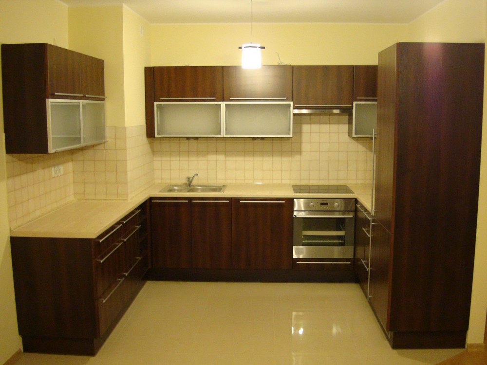 Kompletna zabudowa kuchenna w odcieniach brązu, z beżowymi płytkami i blatem, oświetlona wiszącą lampą z białym kloszem.