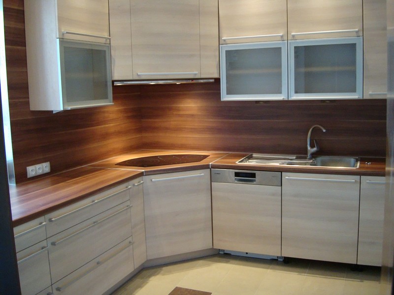 Jasne, nowoczesne meble kuchenne z drewnianym blatem i frontami w odcieniach beżu, uzupełnione o szklane witryny i stalowy zlew.