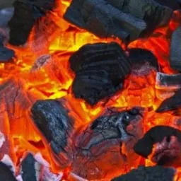 Żarzące się bryły węgla o nieregularnych kształtach, z widocznymi pęknięciami i strukturą, emitujące intensywne pomarańczowe światło.