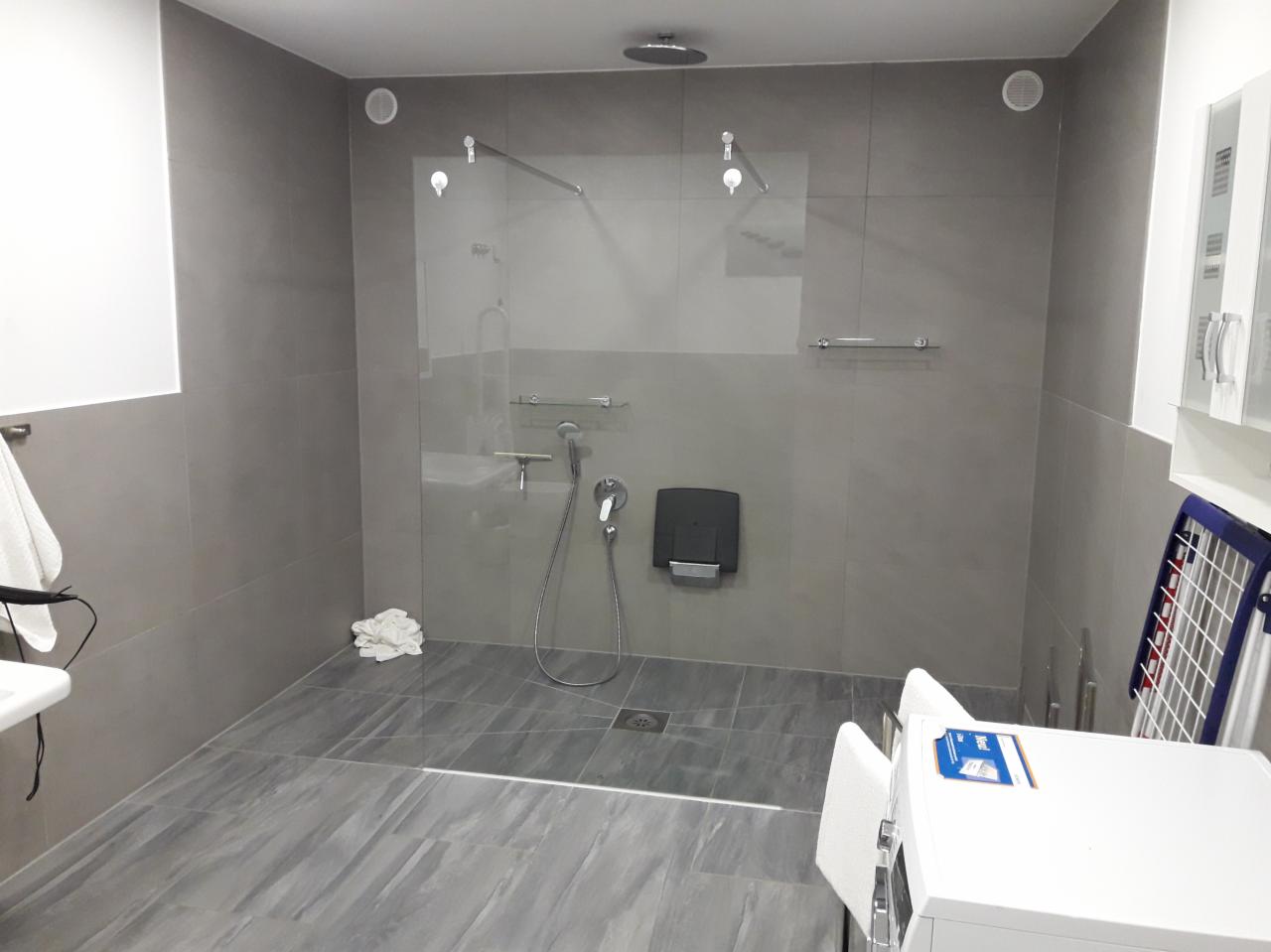 Nowoczesna szara łazienka z kabiną prysznicową typu walk-in, wykończona płytkami imitującymi drewno, z widocznym odpływem liniowym i akcesoriami łazienkowymi.