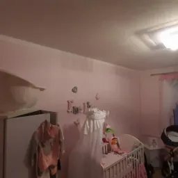 R&oacute;żowe ściany w pokoju dziecięcym z ł&oacute;żeczkiem, w&oacute;zkiem i szafą, na kt&oacute;rej stoi dziecięca wanienka. Świecąca lampa sufitowa oświetla wnętrze.