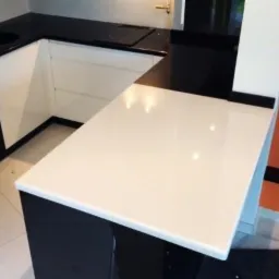 Biały blat kamienny na wyspie kuchennej z czarnymi szafkami, widoczne łączenie blatu z pionową ścianką, płytki podłogowe w tle.