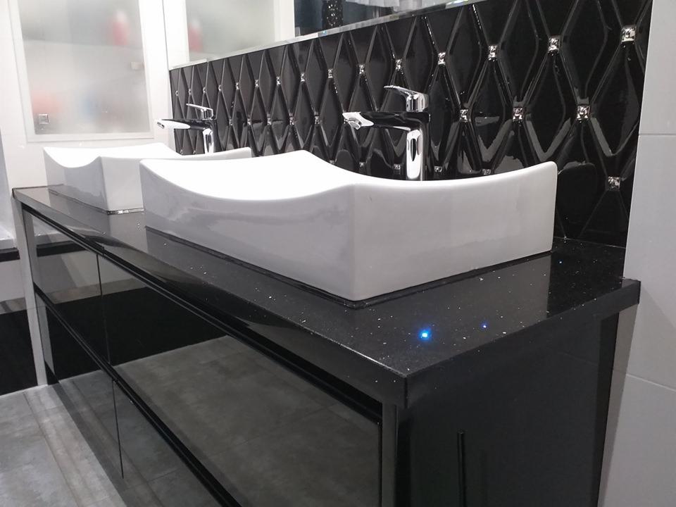 Łazienka z dwoma białymi, nowoczesnymi umywalkami na czarnym, błyszczącym blacie, uzupełniona czarną, diamentową mozaiką z błyszczącymi akcentami.