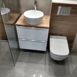 Nowoczesna łazienka z podwieszaną umywalką na drewnianym blacie, szafką z szufladami, podwieszanym WC i szklaną kabiną prysznicową, wykończona w szarościach i drewnie.