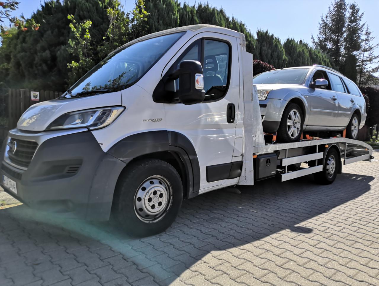 Srebrny samochód kombi zabezpieczony pasami na platformie białej lawety, zaparkowanej na brukowanej nawierzchni, z zielenią w tle.