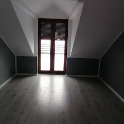 Poddasze po remoncie: szara ściana, drewniana podłoga, okno w skosie. Widok na wykończone wnętrze, ukazujący profesjonalizm prac remontowych.