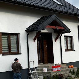 firma remontowo-wykończeniowa - Elewacja domu po malowaniu z widocznym pracownikiem, drabiną i narzędziami. Część ściany w kolorze szarym, reszta biała. Na schodach dynie i farby.