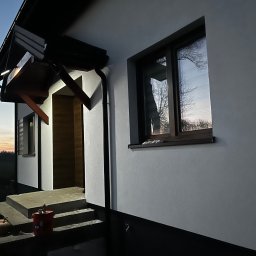 firma remontowo-wykończeniowa - Elewacja domu jednorodzinnego z oknem, po zmroku. Widoczne detale wykończenia, schody wejściowe i puszki po materiałach budowlanych.