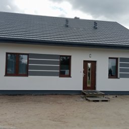 firma remontowo-wykończeniowa - Nowoczesny dom jednorodzinny z szarą dachówką, białymi ścianami z szarymi pasami i drewnianą stolarką okienną, ukazany w pochmurny dzień.