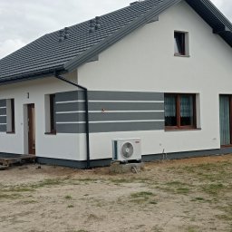 firma remontowo-wykończeniowa - Wykończony dom z białą elewacją i szarymi pasami dekoracyjnymi. Widoczny klimatyzator zewnętrzny, okna z brązowymi ramami i szara dachówka.