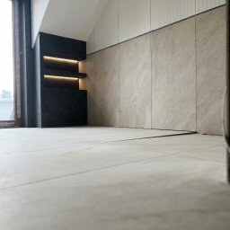 firma remontowo-wykończeniowa - Wnętrze pokoju z wykończonymi szarymi płytkami na ścianie do połowy wysokości i podświetlaną, ciemną wnęką z półkami, z widokiem na okno.