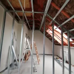 Stelaż z metalowych profili na poddaszu przygotowany do montażu płyt. Widoczne izolacja, okno dachowe, drabiny i elementy konstrukcyjne.