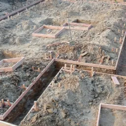 Przygotowanie terenu pod fundamenty: wykopane rowy z drewnianym szalunkiem i pionowymi wspornikami, widoczna ziemia i narzędzia, teren budowy w trakcie prac.