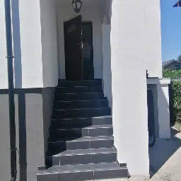 Elewacja domu z białego tynku z ciemnymi schodami wejściowymi wyłożonymi płytkami, oświetlona dekoracyjną lampą wiszącą w łukowym wejściu.