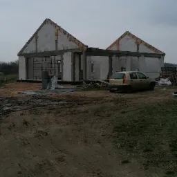 Wznoszenie ścian domu murowanego z betonu komórkowego, widoczne drewniane elementy konstrukcji dachu i zaparkowany samochód osobowy na tle błotnistego terenu.