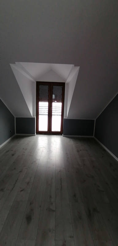 Poddasze po remoncie: szara ściana, drewniana podłoga, okno w skosie. Widok na wykończone wnętrze, ukazujący profesjonalizm prac remontowych.