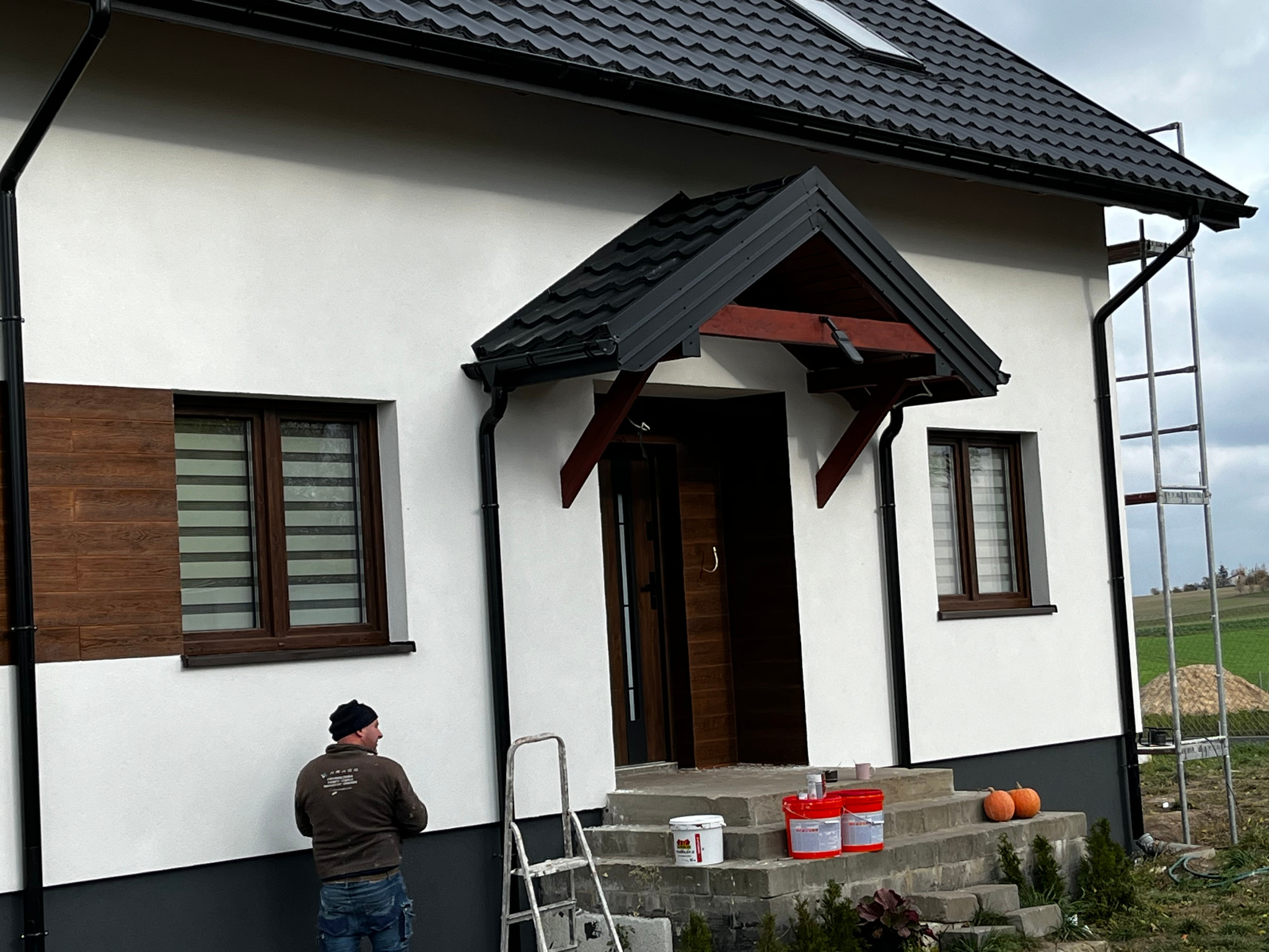 Elewacja domu po malowaniu z widocznym pracownikiem, drabiną i narzędziami. Część ściany w kolorze szarym, reszta biała. Na schodach dynie i farby.