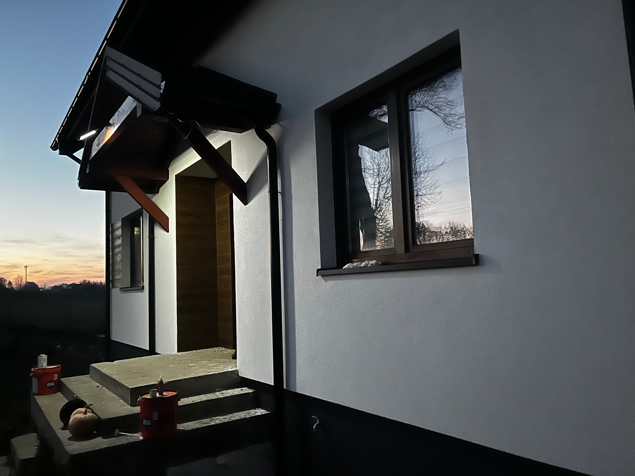 Elewacja domu jednorodzinnego z oknem, po zmroku. Widoczne detale wykończenia, schody wejściowe i puszki po materiałach budowlanych.