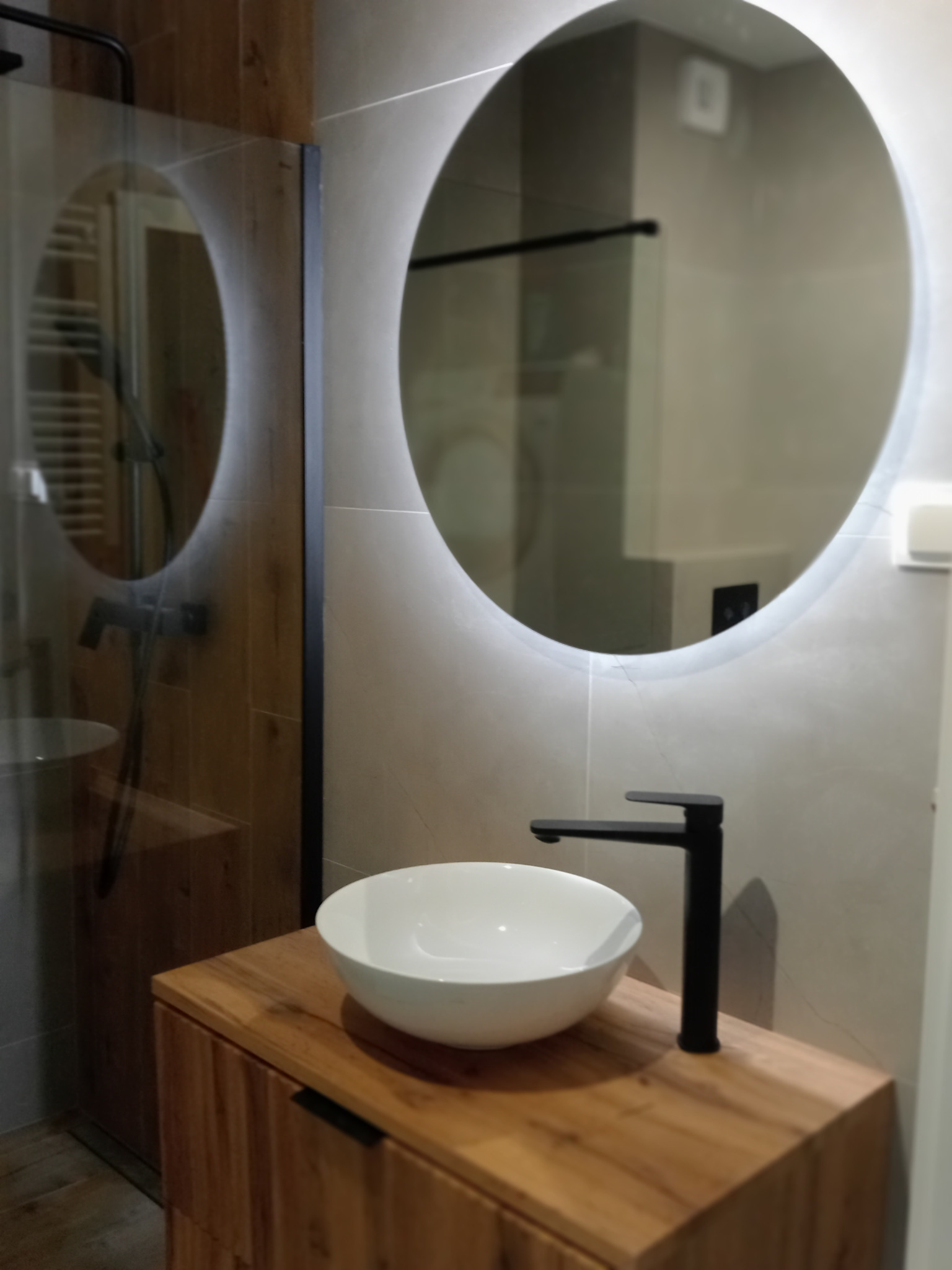 Nowoczesna łazienka z okrągłym lustrem LED, białą umywalką nablatową i czarną armaturą na drewnianej szafce. Widoczna kabina prysznicowa z czarnymi profilami.