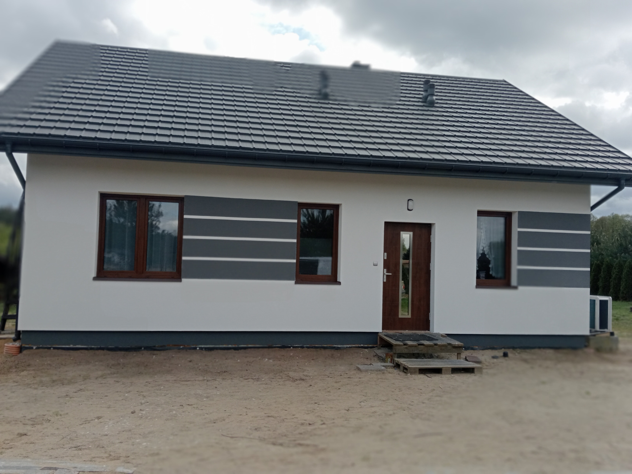 Nowoczesny dom jednorodzinny z szarą dachówką, białymi ścianami z szarymi pasami i drewnianą stolarką okienną, ukazany w pochmurny dzień.