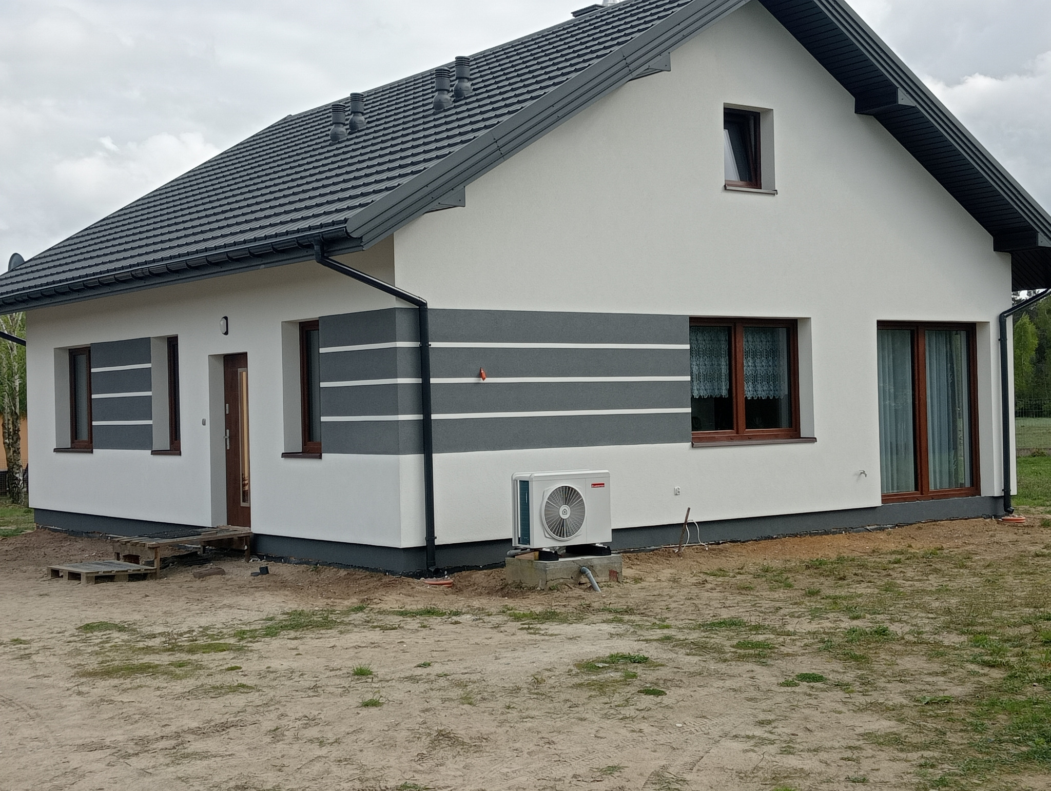 Wykończony dom z białą elewacją i szarymi pasami dekoracyjnymi. Widoczny klimatyzator zewnętrzny, okna z brązowymi ramami i szara dachówka.