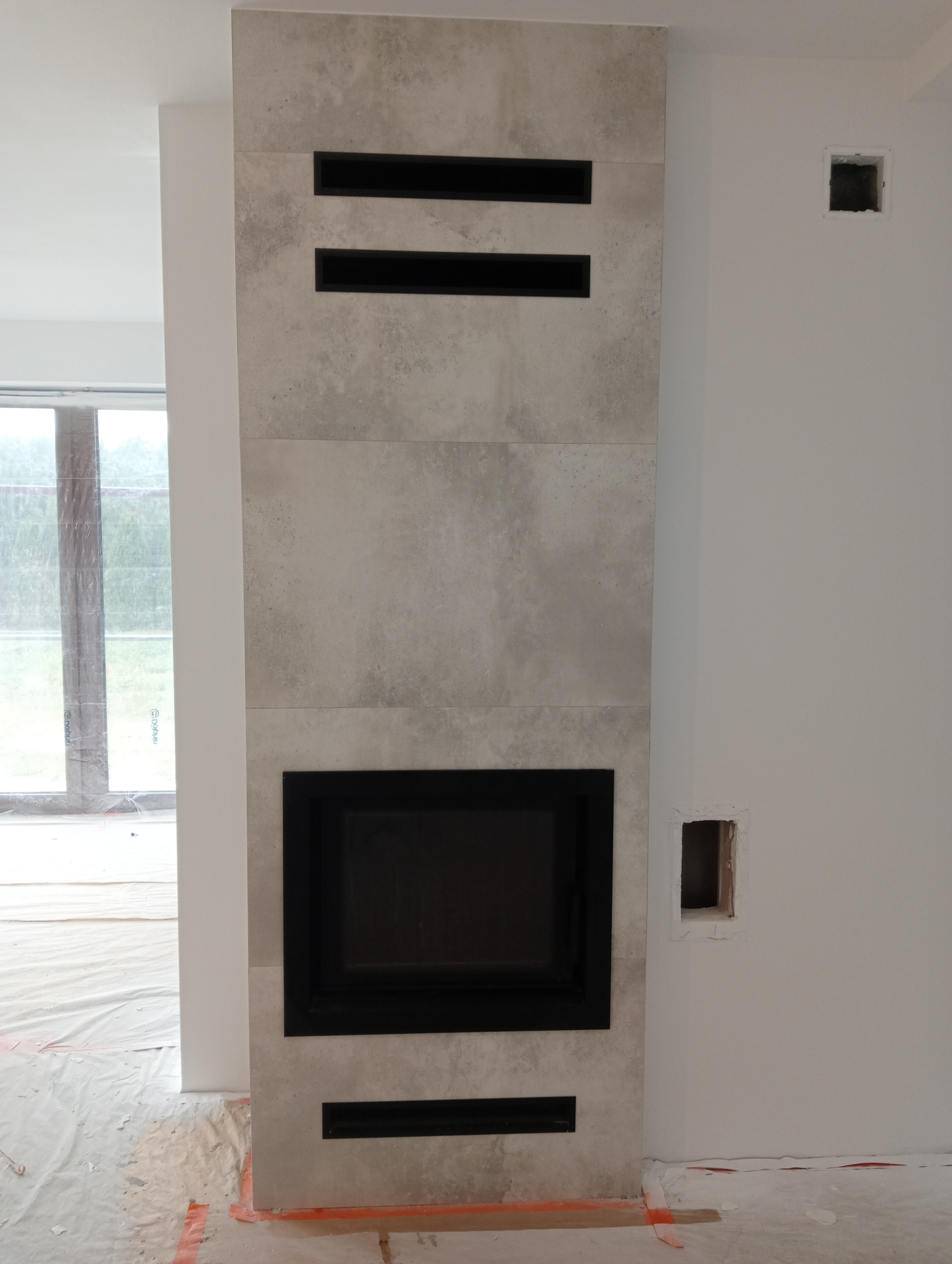 Wykończony kominek z szarego kamienia z czarnym wkładem, widoczne otwory wentylacyjne. Podłoga zabezpieczona folią malarską, ściany białe, surowe wykończenie.