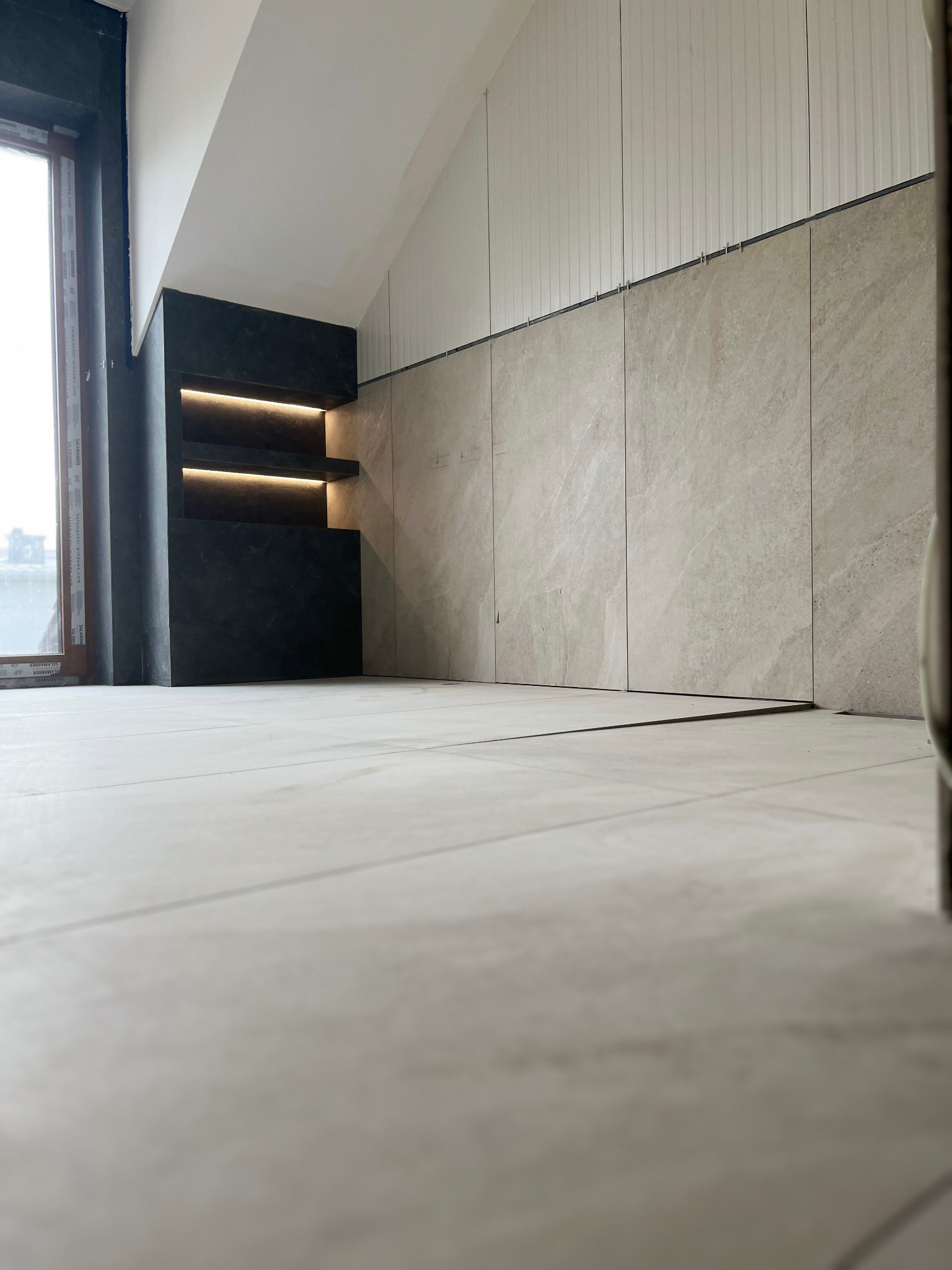 Wnętrze pokoju z wykończonymi szarymi płytkami na ścianie do połowy wysokości i podświetlaną, ciemną wnęką z półkami, z widokiem na okno.